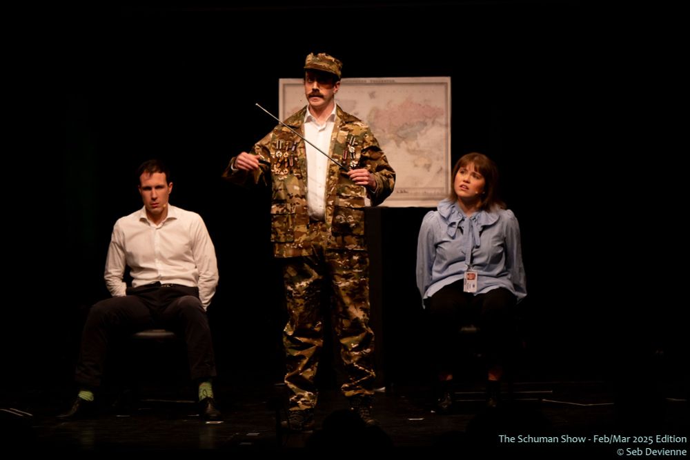 Two actors on stage sitting down with their hands tied behind their backs, in office wear. In between them, another actor dressed up as a general of some sort, in camo wear.