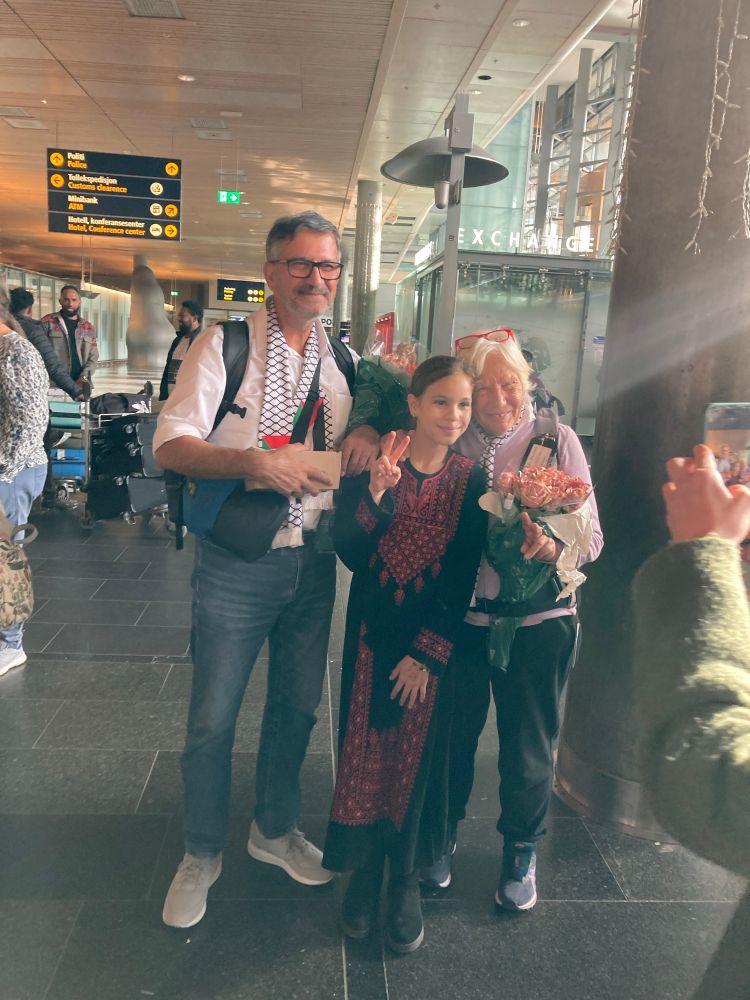 En godt voksen mann og en godt voksen dame står med blomster i armene og palestina-skjerf rundt halsen på en flyplass. De smiler. Mellom dem står et barn i palestinsk tradisjonell drakt. De viser V-tegnet.