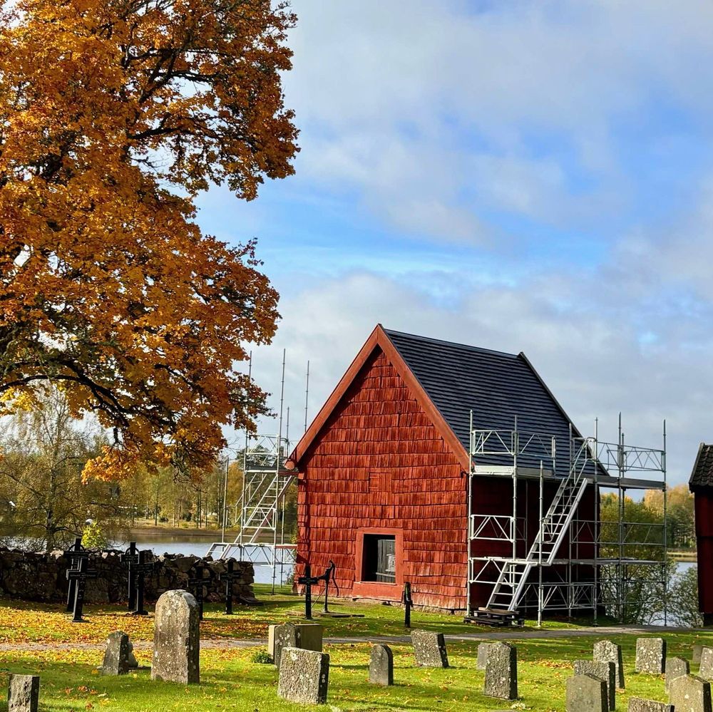 Ett solbelyst litet falurött förrådshus i utkanten av en kyrkogård med ett stort träd i höstfärger i vänsterkanten och en sjö i bakgrunden.