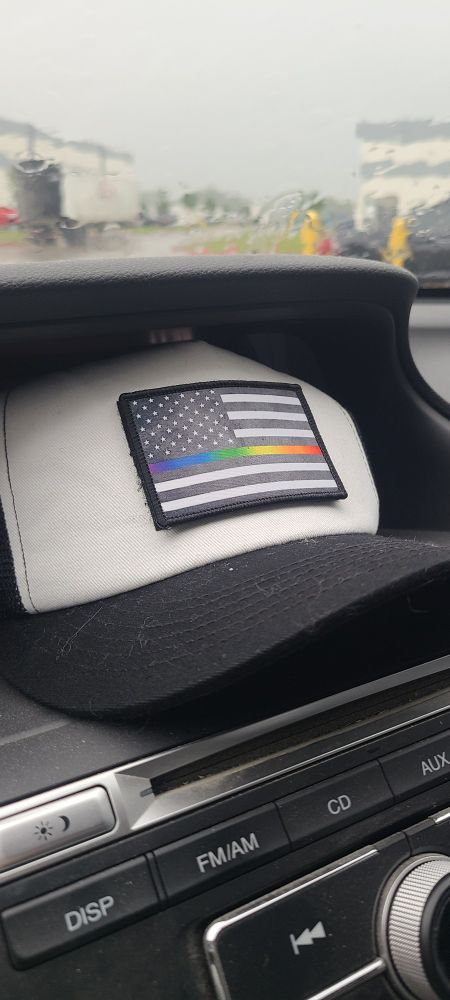 A black brimmed, white front, trucker hat. The original Black American flag has been removed, and replaced with a black and white American Flag, where the 8th stripe has been replaced by a rainbow gradient. The poster call this "The thin Gay line" hat. 