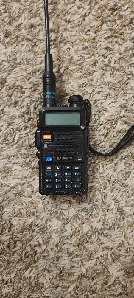 A Baofeng UV5R handheld radio laying on carpet. It has a Long antenna installed
