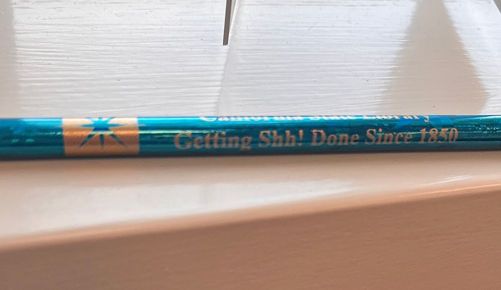 A picture of “merch” from the California State Library. Picture is a side view of a teal holographic pencil with the text: California State Library Getting Shh! Done Since 1850
