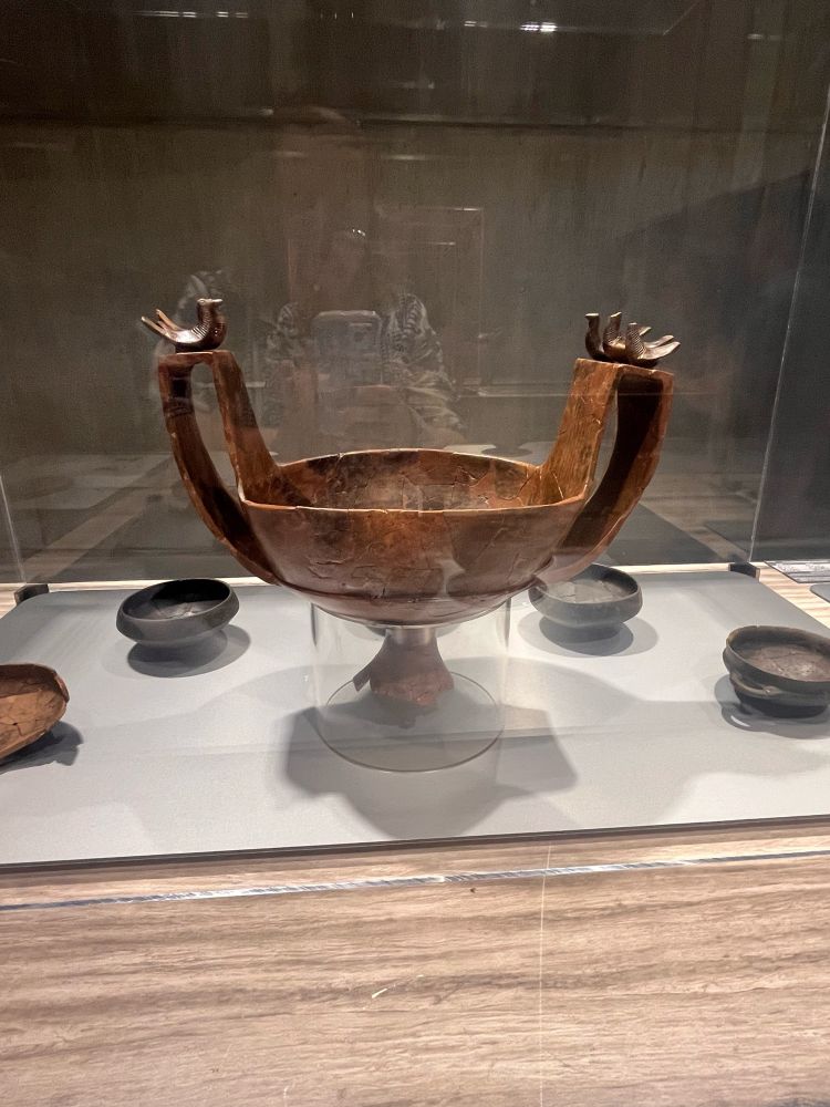 Bronze vessel with tall, curved handles ending in stylized human hands, displayed in a glass case at the Rocca di Frassinello Etruscan Museum. The bowl rests on a pedestal base, surrounded by smaller blackware cups.