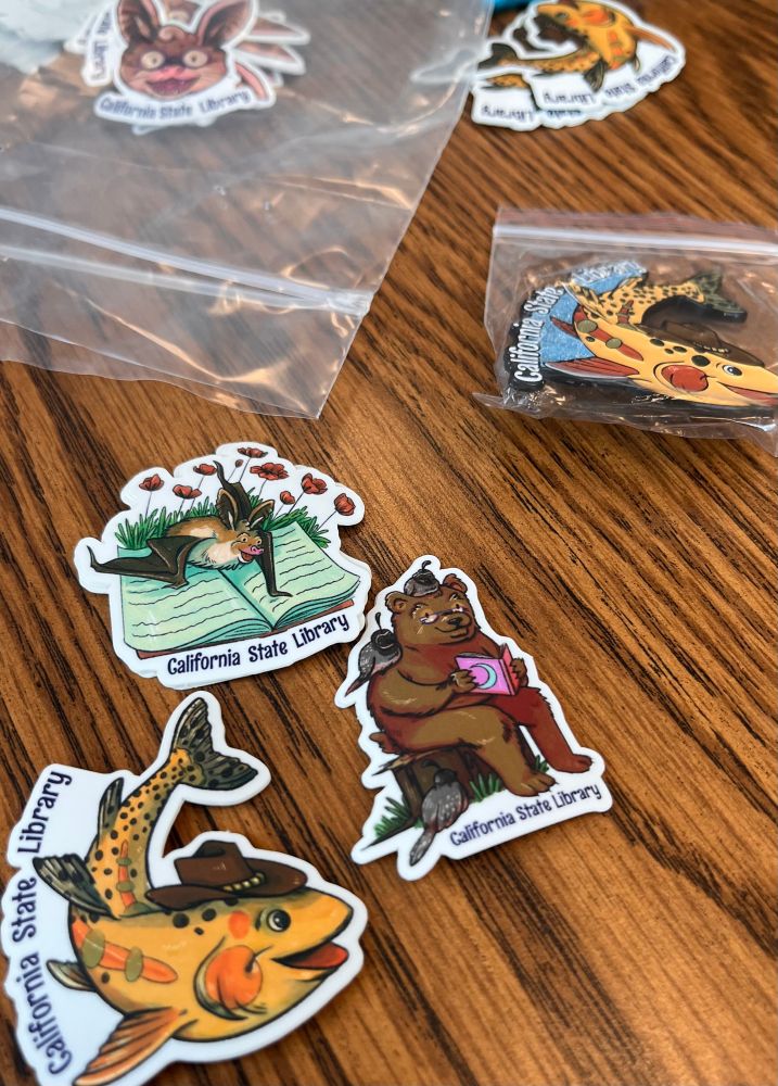 More merch from the state library. In the front of the picture are three state symbol stickers, illustrations by Megan Porch. To the left is a golden trout wearing a cowboy hat. To the right is a grizzly bear rearing a book to three quails (including one on top of their head). Above that is a sticker featuring a pallid bat in an opened book with golden poppies behind them. 