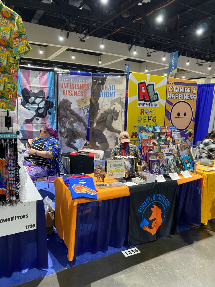 A colorful vendor booth at San Diego Comic-Con featuring the Forester Universe by Tim Susman (Kyell Gold). Prominently displayed are vertical banners for two novels, “Unfinished Business” and “Dead Right,” each featuring anthropomorphic wolf characters. Behind the table hangs a large trans pride flag with a pawprint. The table is covered in orange and blue cloth with the Forester Universe fox-and-book logo, and displays a variety of books, shirts, and signs for eBooks and audiobooks. Two people in patterned shirts sit behind the booth, and adjacent booths are partially visible. Booth number 1236.