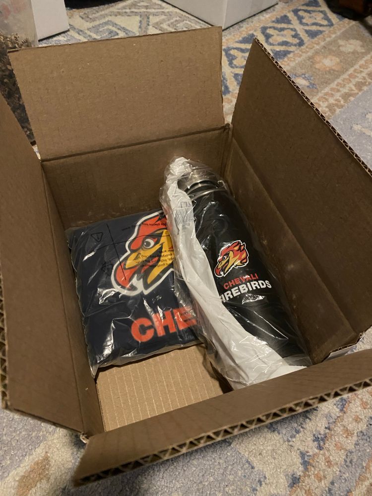 Open cardboard box containing a Chevali Firebirds T-shirt and black water bottle wrapped in plastic.