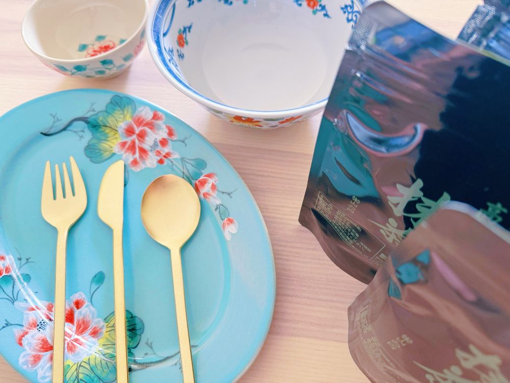 水色に花柄模様のオーバル皿、ゴールドのカトラリーセット、ベージュに花柄のお椀と白地に中華風の絵柄の入った丼、パック入り抹茶がテーブルに並んでいるところ。