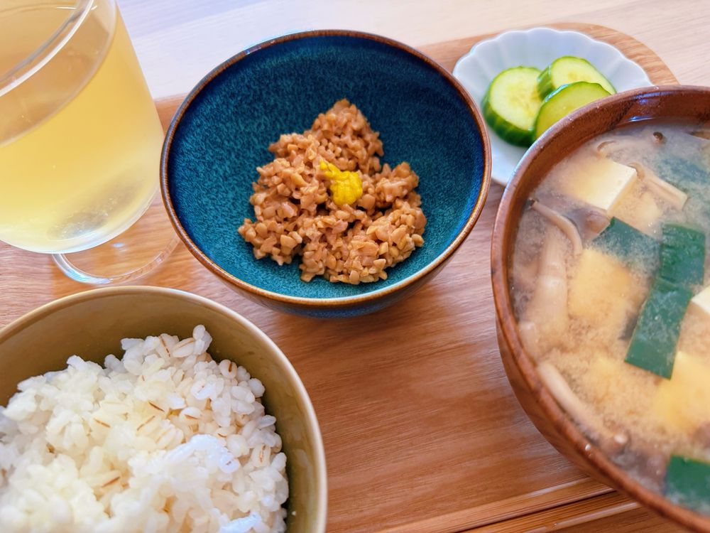 長方形のお盆に乗ったお昼ごはん。左前から時計回りに麦の入った白米、グラスに注がれた黒豆茶、ひきわり納豆、きゅうりの浅漬け、具沢山のお味噌汁。