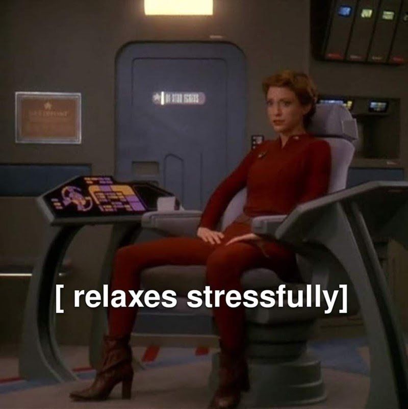 Kira Nerys wearing a Maroon Bajoran uniforming sitting in the Captain's seat of the Defiant with an anxious facial expression. Text: [relaxes stressfully]