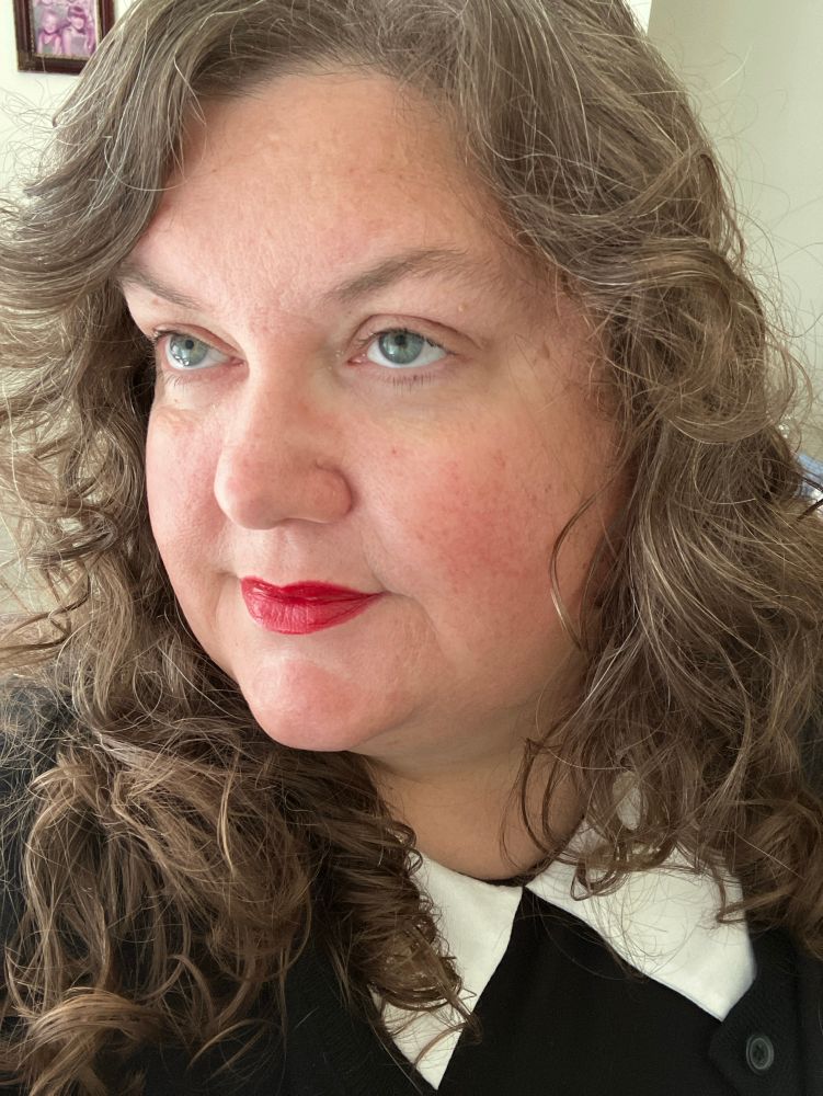 femme in a black dress with a pointed white collar. Their long medium auburn hair is curly and cascades down their shoulders. They have green eyes and are wearing red lipstick. They are looking nightly out of frame.