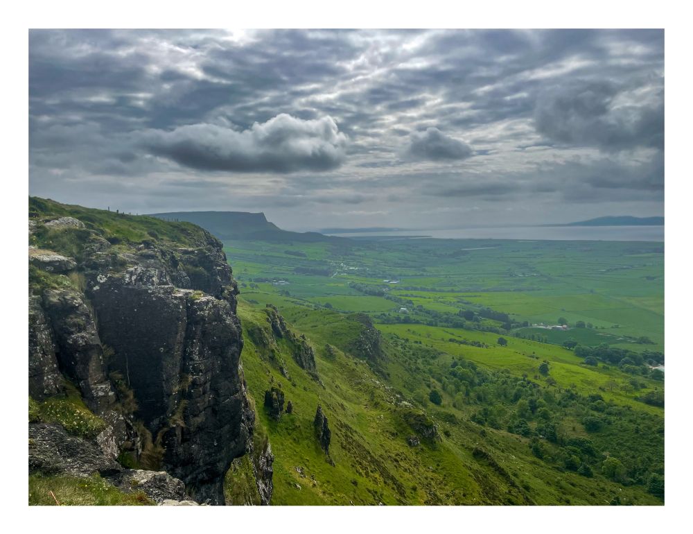 A dramatic landscape featuring a rugged cliff edge in the foreground, overlooking a vast expanse of green fields and rolling hills. The sky is overcast with moody, textured clouds casting diffused light across the scene. In the distance, a flat-topped mountain rises against the horizon, and beyond that, a glimpse of the sea is visible under the cloud-covered sky. The overall atmosphere is serene and wild, evoking the beauty of untamed countryside.