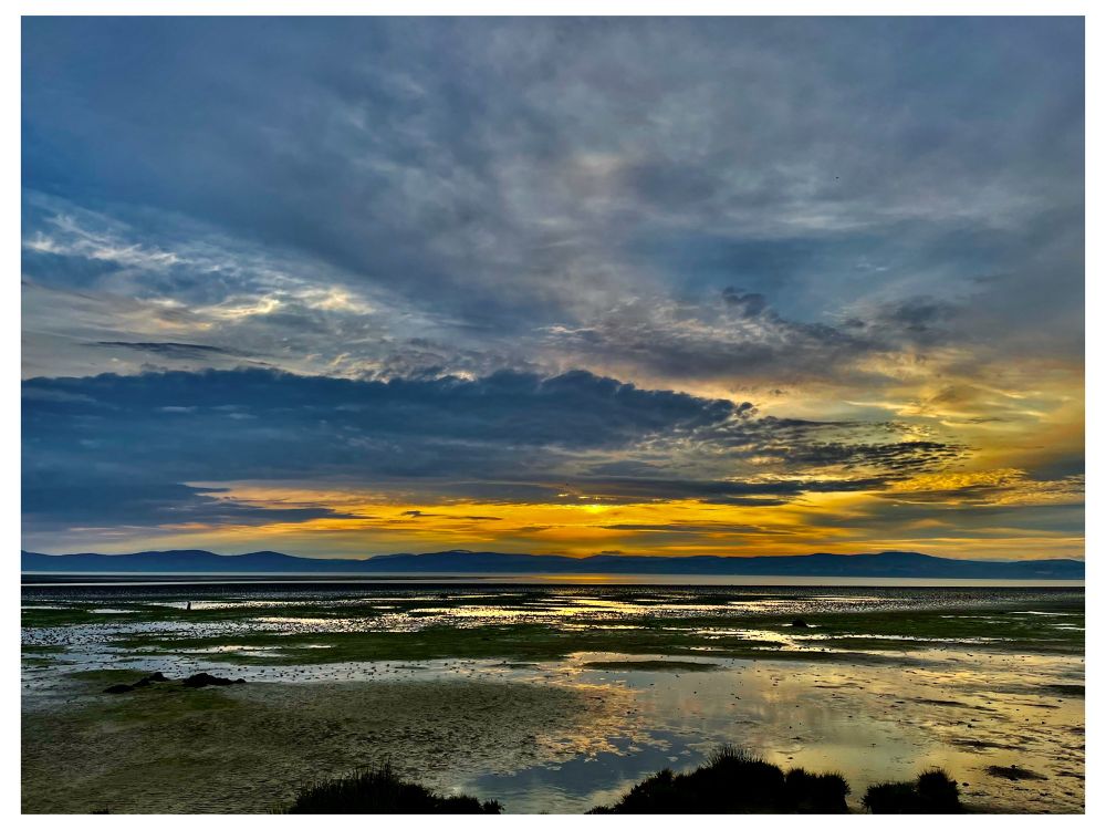A dramatic coastal sunset scene with a wide expanse of shallow tidal pools in the foreground, reflecting the vibrant colors of the sky. The sky is filled with layered clouds in varying shades of blue, grey, and gold, with a burst of intense yellow-orange light near the horizon. Distant hills or mountains create a dark silhouette against the glowing sky, and patches of green seaweed and grass are visible in the wet sand and water below.
