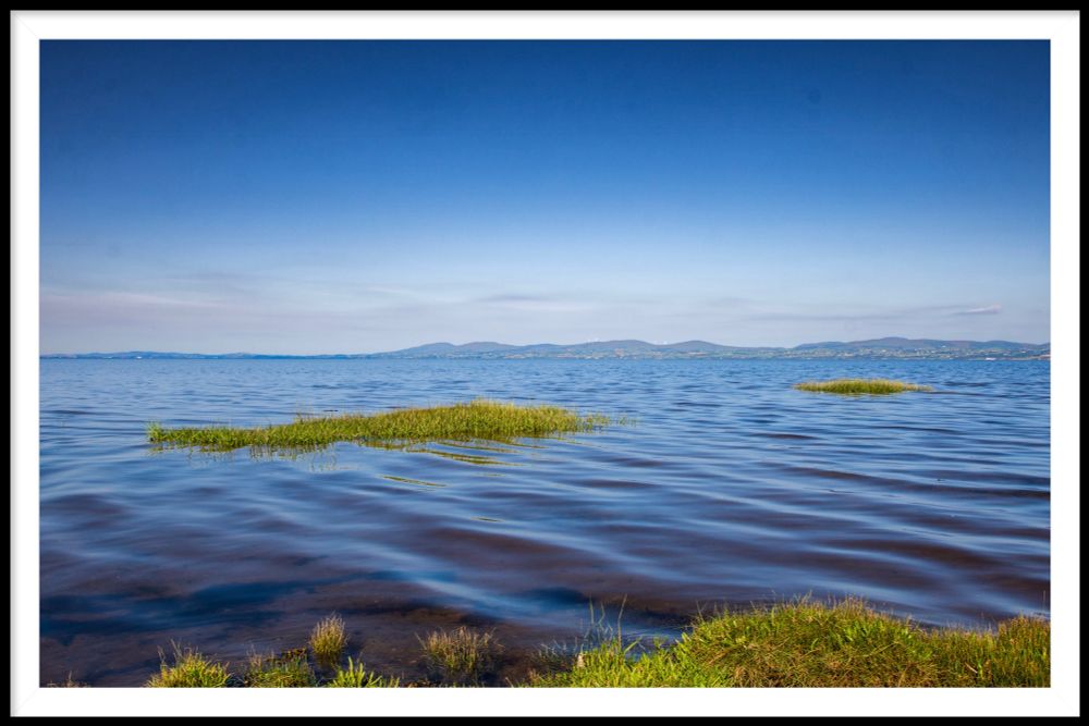 On a hot summer’s day. Lough Foyle ripples towards me. Blue skies and blue waters. Green tufts of grass are visible. Mountains of Donegal behind. 