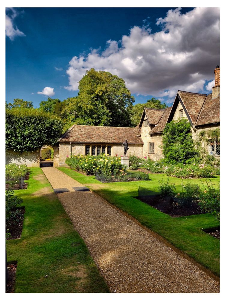 A gravel path leads through a neatly maintained garden with lush green lawns and flower beds filled with blooming roses. The path ends at a charming stone building with steeply pitched tiled roofs and mullioned windows. A statue stands prominently in front of the building, surrounded by yellow and pink flowers. To the left, an arched ivy-covered gateway opens to another area. Tall trees and a vibrant blue sky with dramatic white and gray clouds complete the idyllic scene.