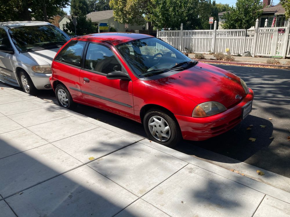 A bright red Chevy Metro parked along the sidewalk on a residential street. 