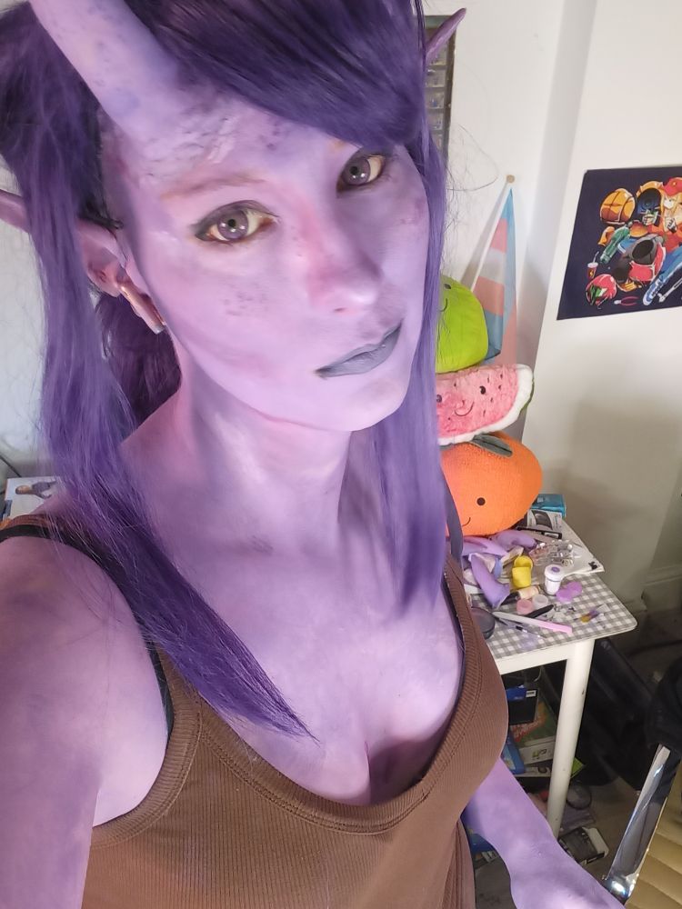 A photo of BisectedBrioche dressed as a purple tiefling. She has purple freckled skin, claws, sharp teeth and horns. Her ear is pointed and has three rings on the outer edge. She is wearing a brown tank top. She is looking into the camera from below.