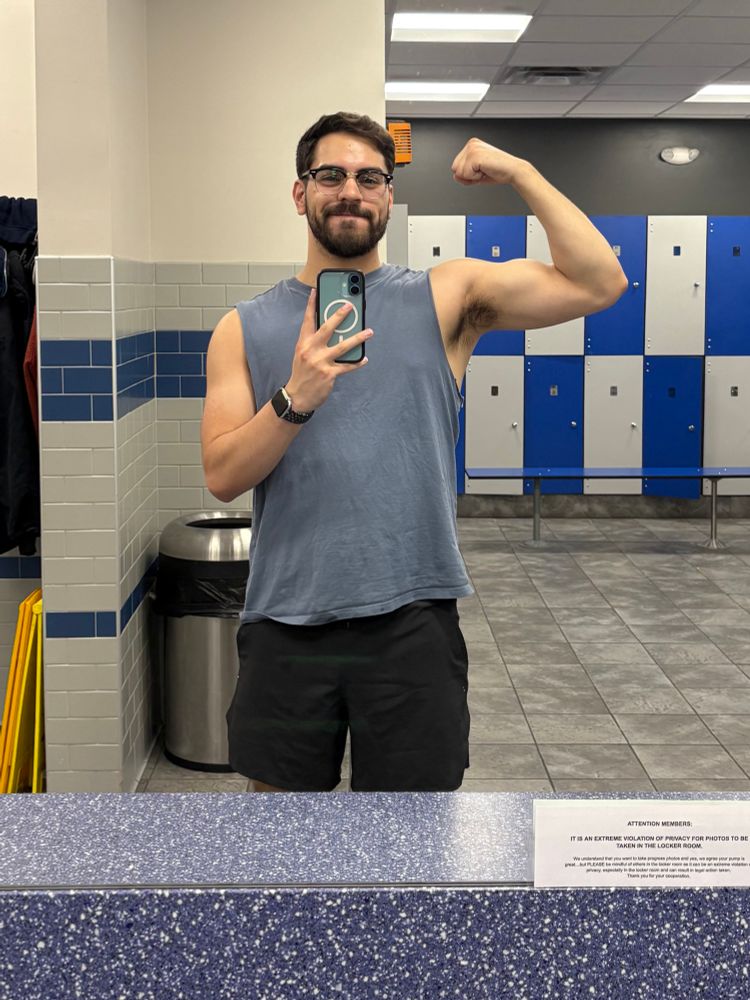 A man taking a photo in the gym locker room mirror. He’s flexing one bicep while wearing a blue tank top
