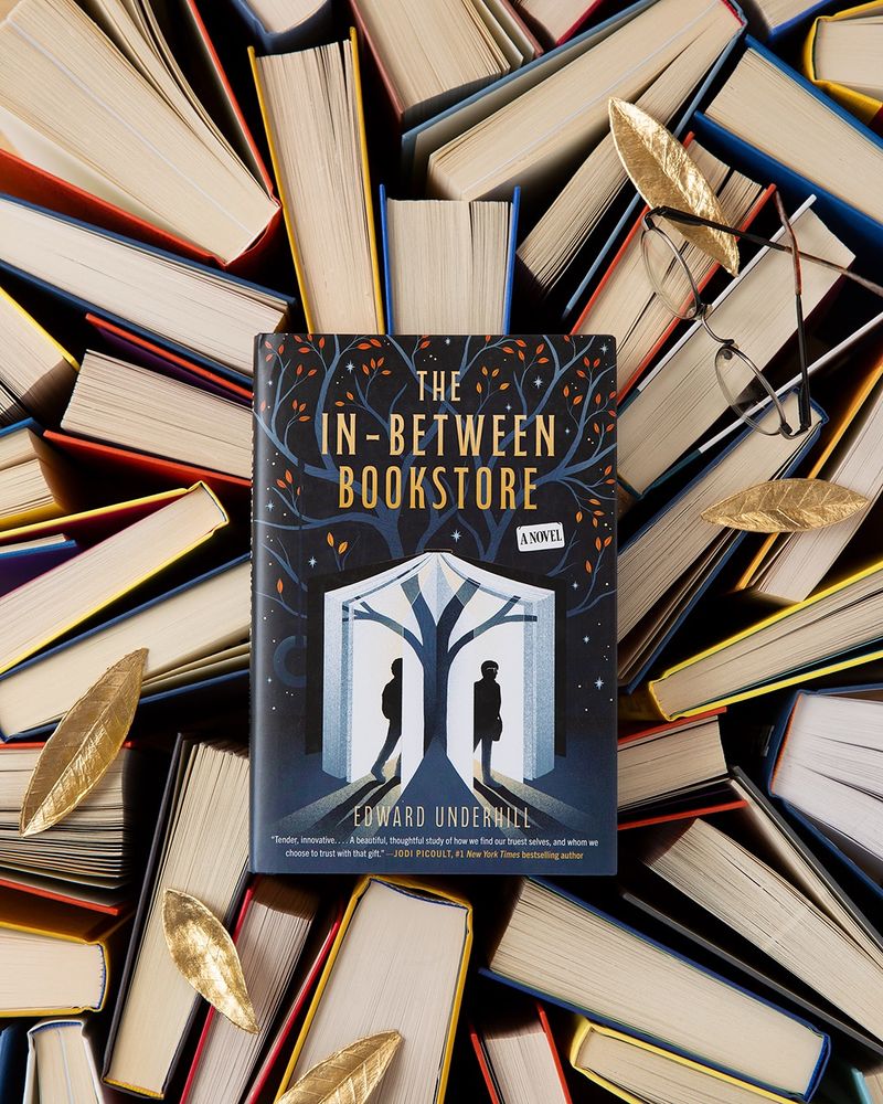 A hardcover copy of The In-Between Bookstore, which is blue and features a tree growing out of the pages of a book, and two silhouettes walking into and out of the book, against a backdrop of other books, leaves, and a pair of glasses.