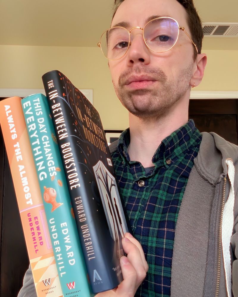 The author, a white trans man with glasses, holding the books he has written: Always the Almost, This Day Changes Everything, and The In-Between Bookstore