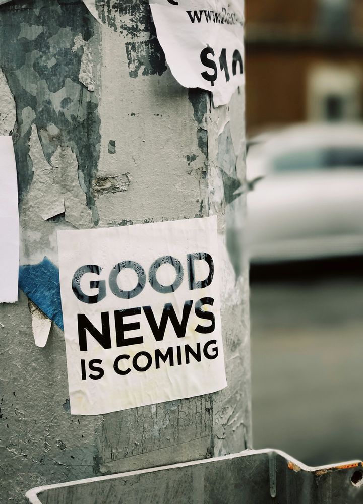 A weathered utility pole stands tall in an urban setting, its surface adorned with several torn and partially visible posters. The most prominent poster, positioned at the center, boldly declares "GOOD NEWS IS COMING" in striking black and yellow letters. The background is slightly blurred, revealing a parked car and buildings, hinting at a bustling city environment. The overall scene evokes a sense of anticipation and curiosity, as if the message on the poster is a beacon of hope amidst the everyday hustle and bustle. Photo by Jon Tyson on Unsplash