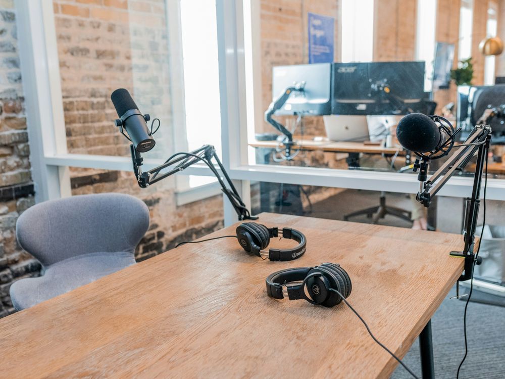 A cozy and modern podcast recording studio is set up with a wooden table at the center. On the table, there are two pairs of sleek black over-ear headphones, ready for use. Two adjustable microphone arms are attached to the table, each holding a professional-grade microphone, poised for recording. To the left, an empty gray chair with a stylish curved backrest invites the next guest. In the background, through a glass partition, several computer monitors and office equipment can be seen in another room. The room has exposed brick walls and large windows that let in an abundance of natural light, creating a warm and inviting atmosphere perfect for recording engaging audio content like podcasts or interviews. (Photo by Austin Distel on Unsplash)