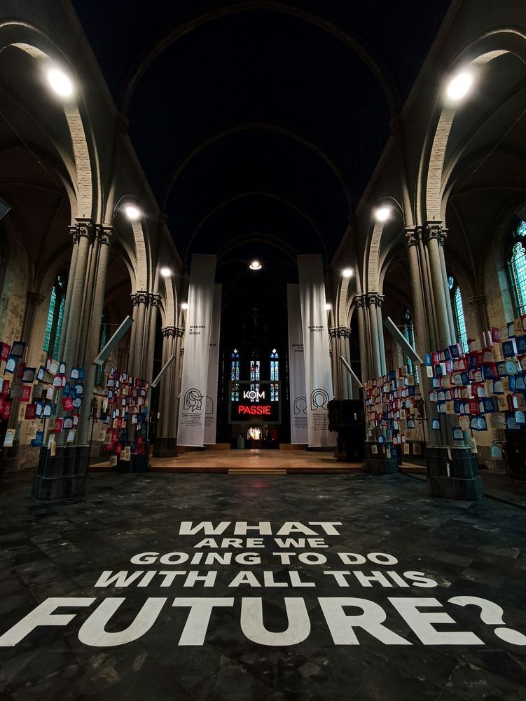 The image captures the majestic interior of a grand, gothic-style church. The high vaulted ceilings and tall, arched windows create a sense of awe and reverence. The floor, made of dark stone tiles, features a striking centerpiece: large white text that boldly asks, "WHAT ARE WE GOING TO DO WITH ALL THIS FUTURE?" This thought-provoking question stands out against the historical backdrop. The walls are adorned with vibrant artwork and banners, adding a splash of color to the otherwise somber tones of the architecture. At the far end of the church, an illuminated sign with the word "PASSIE" in red letters glows warmly, blending the old with the new. The overall atmosphere is a harmonious fusion of historical grandeur and contemporary artistic expression, inviting contemplation and reflection. Photo by Lil Mayer on Unsplash