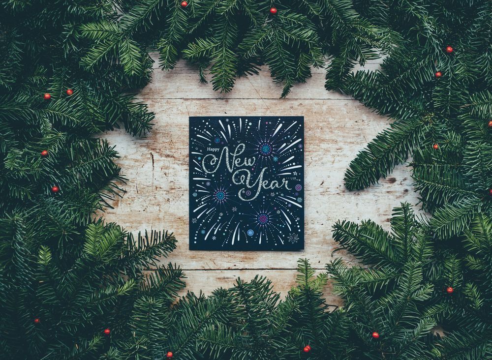 A black, squared, board with the text "Happy New Year" written in silver sparkling text. The text is surrounded by painted fireworks in pink, purple, green, blue and silver. The background is made up by light wooden board with a frame of green pine branches with red berries. Photo by Annie Spratt on Unsplash
