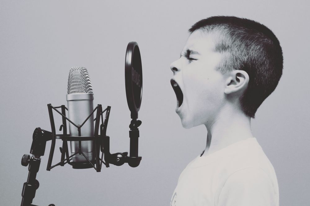 A child screaming into a microphone with a pop-filter in front of it. The picture is a black and white photography taken so that the boy and microphone is viewed from the side. 