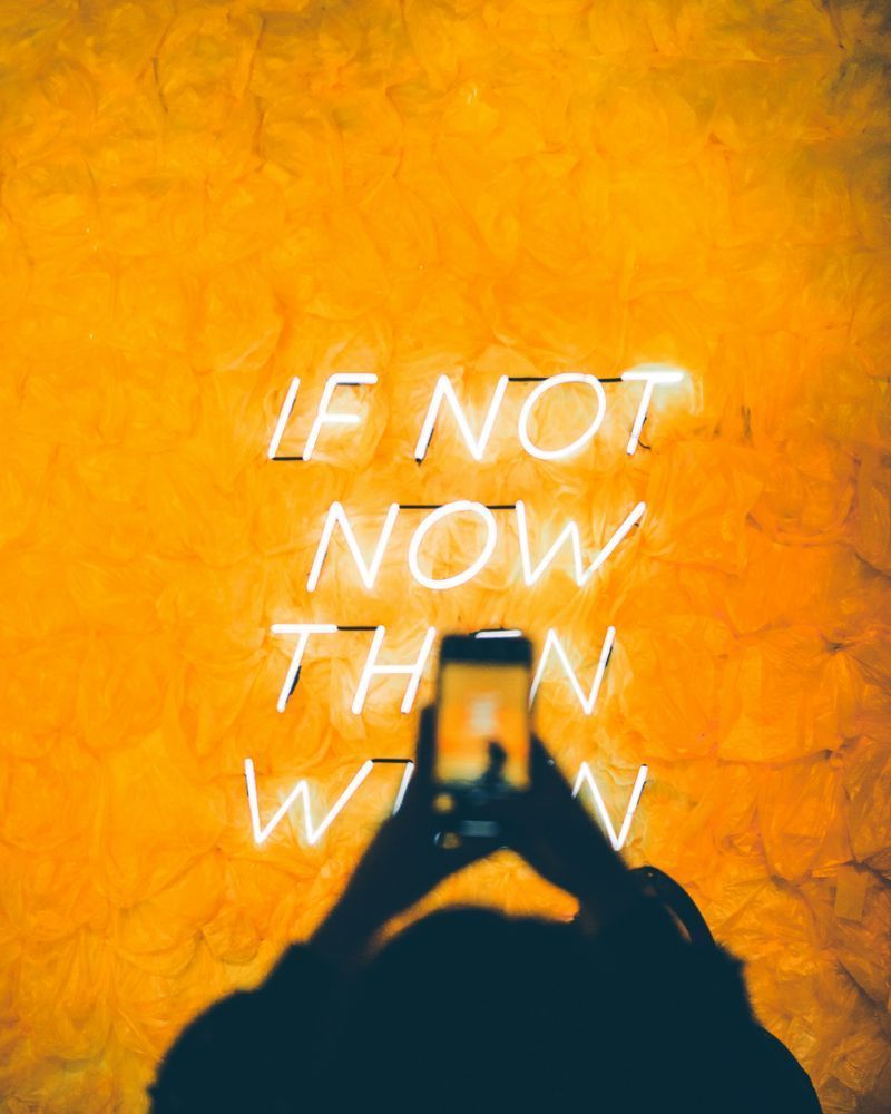 A picture, taken from above, on a orange background with white neon-light letters forming the words: "If not now, then when". In the foreground there is an out of focus phone, taking a picture on the sign, in the hands of a person. Photo by Luis Villasmil on Unsplash