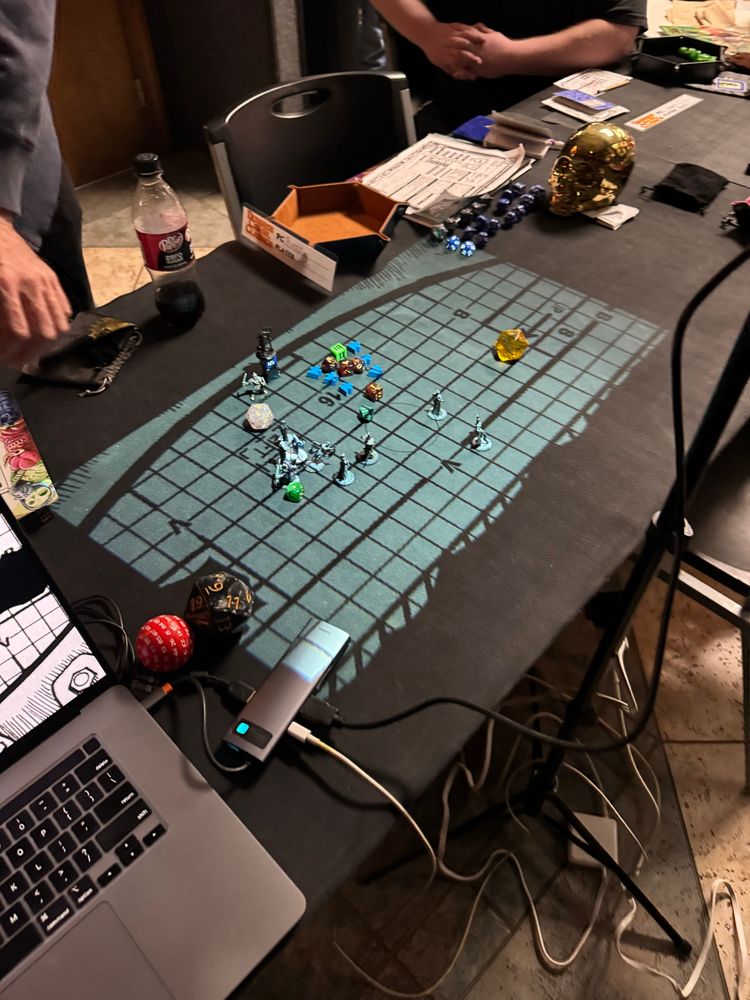 Inside a gaming store on a long table covered in black neoprene mats, a projection of a grid map is displayed from the TTRPG game, Expedition to the Barrier Peaks.

Also on the table is a laptop, a bronze skull various electronic components, character sheets and dice trays.

On the projection itself are a variety of items used for theater of the mind, such as grey miniatures depicting the player’s characters, blue meeples, different d-sided die, a d30, d6, large d20 for the variety of monsters. And a random d100 next to the laptop.