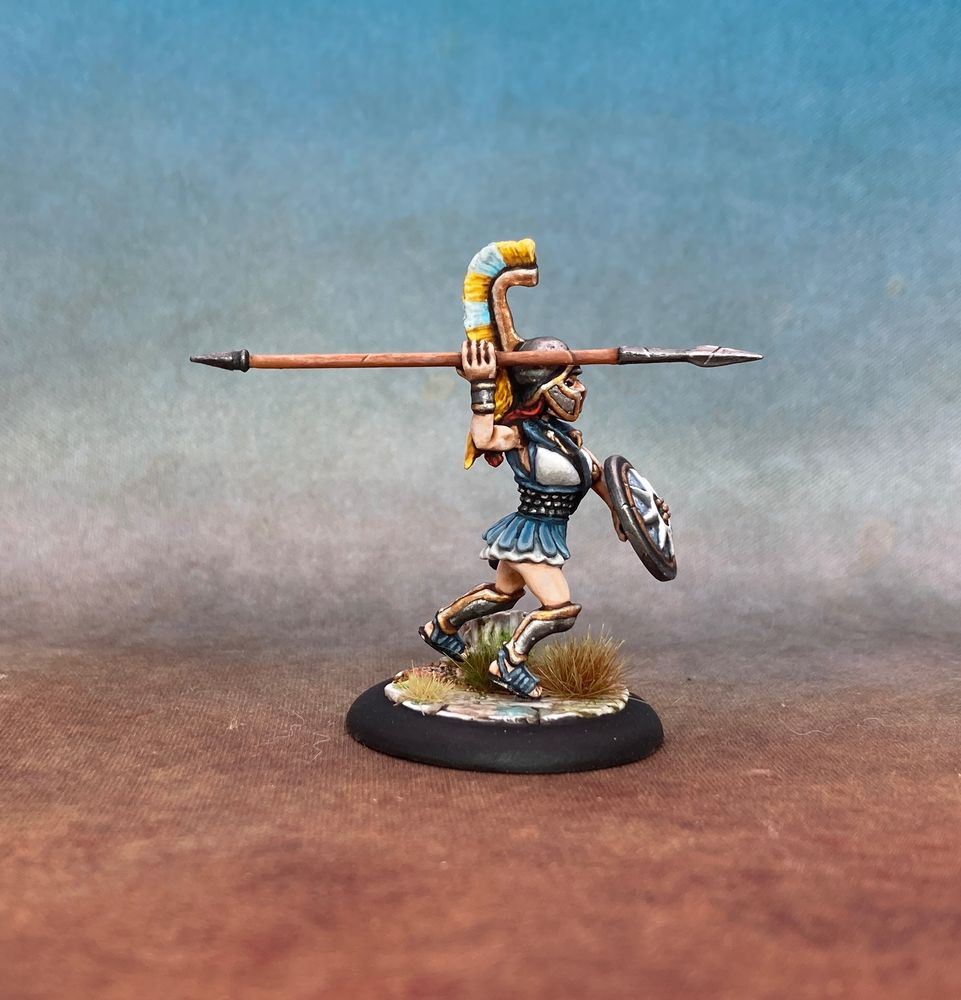 Woman in hoplite panoply charging and yelling. She’s in white, blue grey and pale yellow.