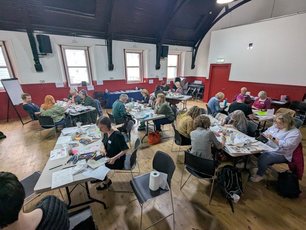 A photo of a room full of people sitting around 6 tables with paper and art materials in front of them. They are painting and chatting, and soft light is coming in from the windows.