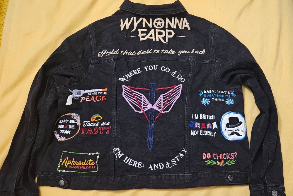 A black denim jacket with Wynonna Earp and ilk embroidered on it against a yellow background. 