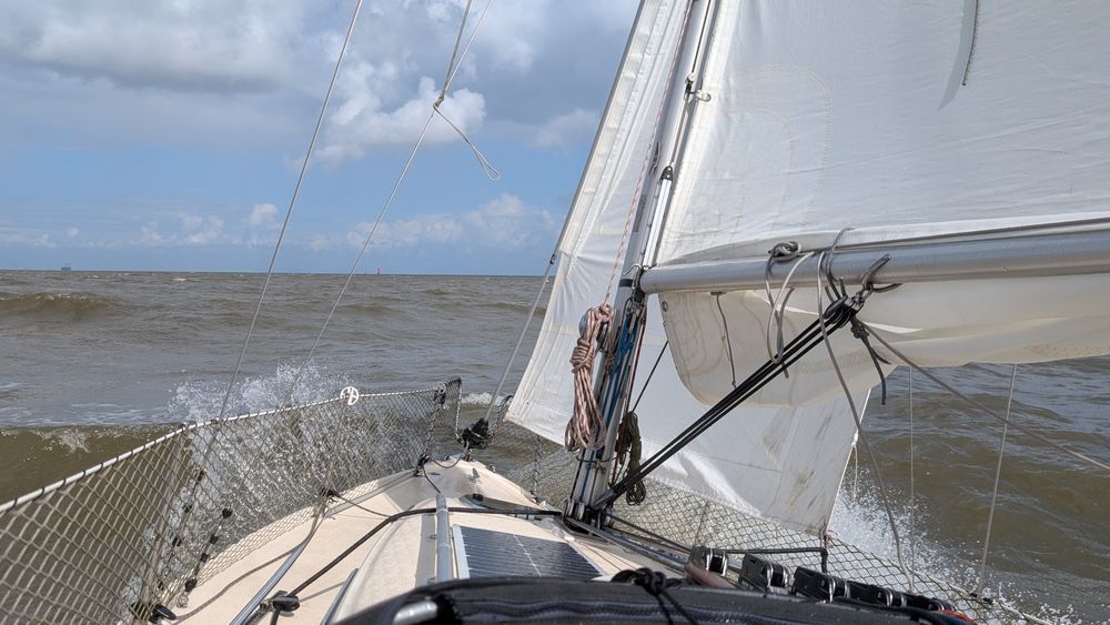 Blick auf das Vorschiff eines Segelbootes auf die Nordsee bei mäßigem Seegang. Das Boot ist leicht zur Seite geneigt, das Großsegel ist dichtgeholt und steht voll im Wind. Gischt spritzt über die Reling, der Himmel ist teils bewölkt, teils blau. Am Horizont ist eine rote Tonne sowie ein Frachtschiff zu erkennen.
