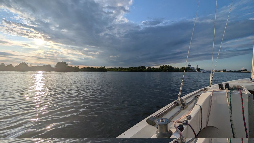Ein Segelboot auf dem Wasser. Das Bild ist im Weitwinkel aufgenommen. Man sieht Wolken und auch ein wenig die Sonne.