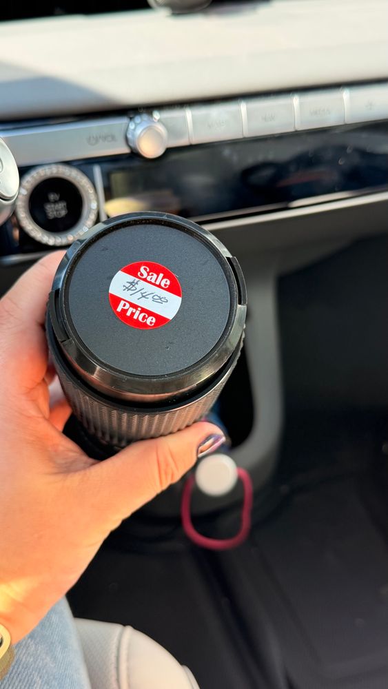 A close-up of the same lens, viewed from the top. A red-and-white sticker on the lens cap reads “Sale Price $14.00” handwritten in black ink. The hand holding it has metallic dark-blue nail polish, and the car interior is visible in soft daylight.