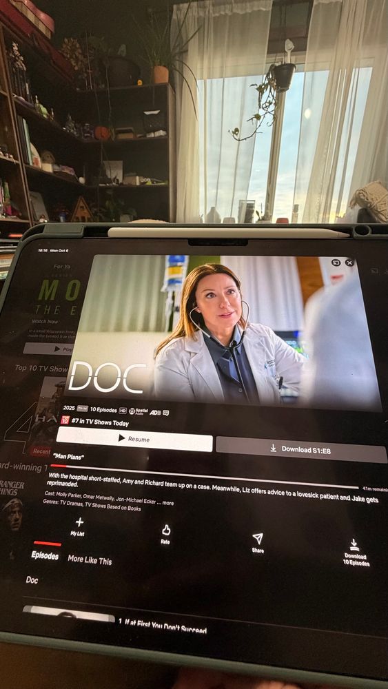 A tablet screen showing the Netflix series Doc, paused on a scene with a female doctor wearing a white coat and stethoscope. The tablet rests in front of a window with sheer curtains, and behind it is a cozy room with shelves full of books, plants, and decorations. The atmosphere feels homely and warm, with afternoon light coming through the window.
