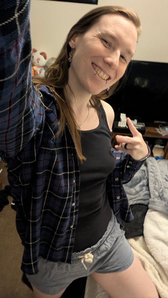 Alice stands taking a selfie while making a peace sign, she is wearing her usual blue flannel, a black tank top, and light blue shorts, in the background is a tv and clutter!