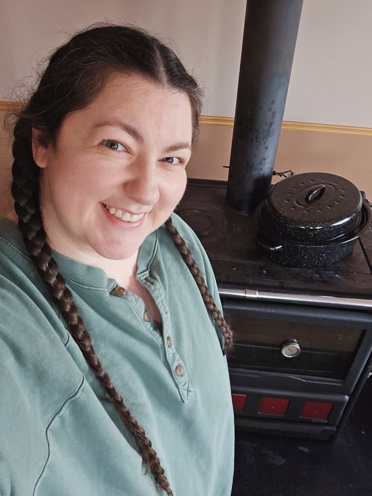 Image is a selfie of Kaylee smiling proudly beside a large black covered pot on her black iron woodstove stove. Kaylee is wearing a green quarter buttoned, oversized sweatshirt. Her hair is in two very long French braids 