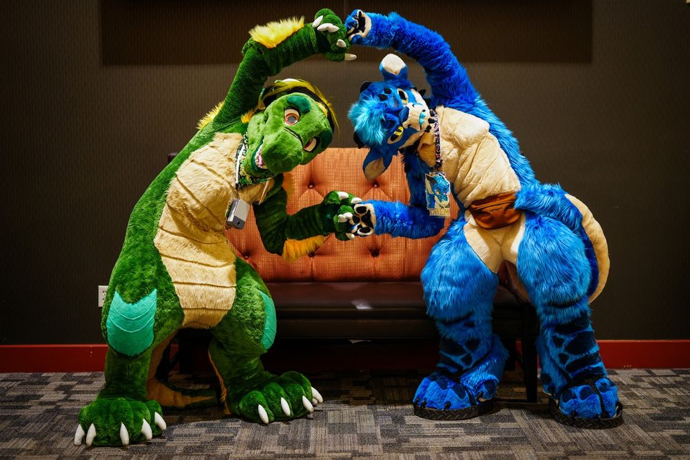 Two people in dragon fursuits, one green and one blue, doing the Dragon Ball Fusion Dance pose with fingers touching.