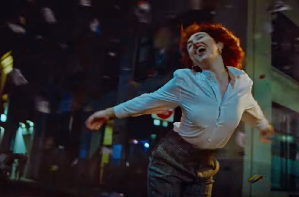 Chappell Roan in her music video for The Subway with a red bob cut and wearing a white blouse and grey dress pants . 