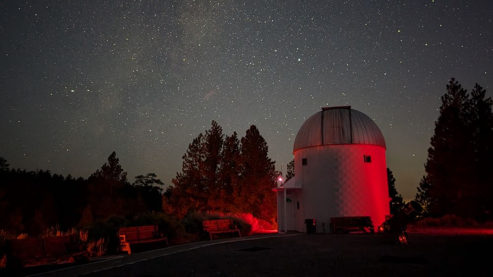 photo of the night sky at Pine Mountain Observatory southeast of Bend, Oregon taken at 4:35am on Sunday, 11 Aug 2024. A white brick building topped with a silver-colored metal telescope dome is bathed in red light, with trees in the background. In the sky can be seen part of the Milky Way, with Lyra seen above the dome and Altair at the top of Aquila on the left side of the image. In front of the telescope building is a large portable Dobsonian telescope on a flat stretch of gravel. 