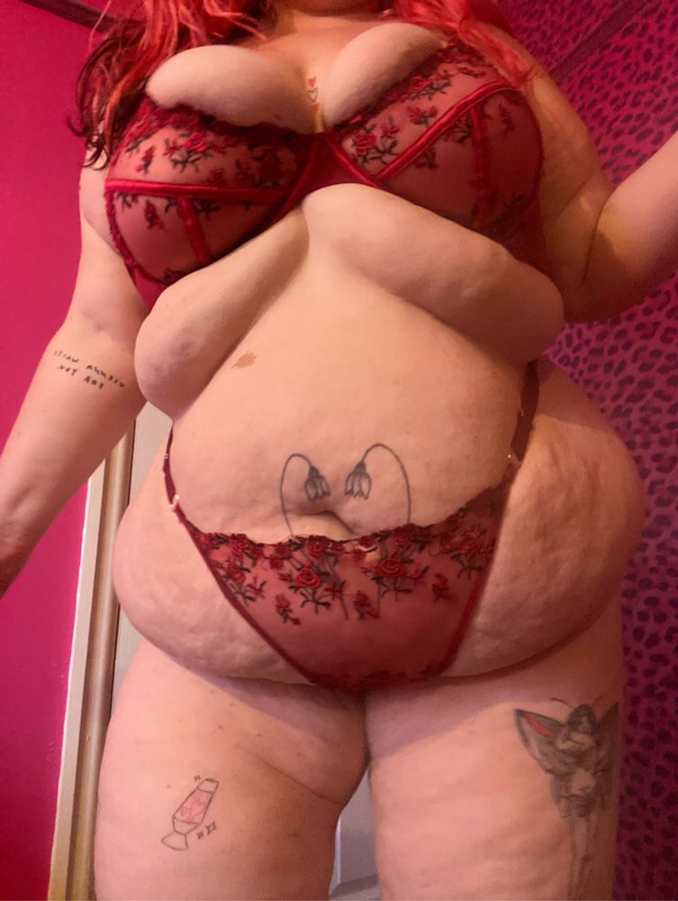 eva wears a red underwear set, matching bra and panties with flowers on, neck to knee shot standing up with legs slightly spread. heart tattoo around her belly button. fairy tattoo on right thigh & lava lamp on left thigh. 