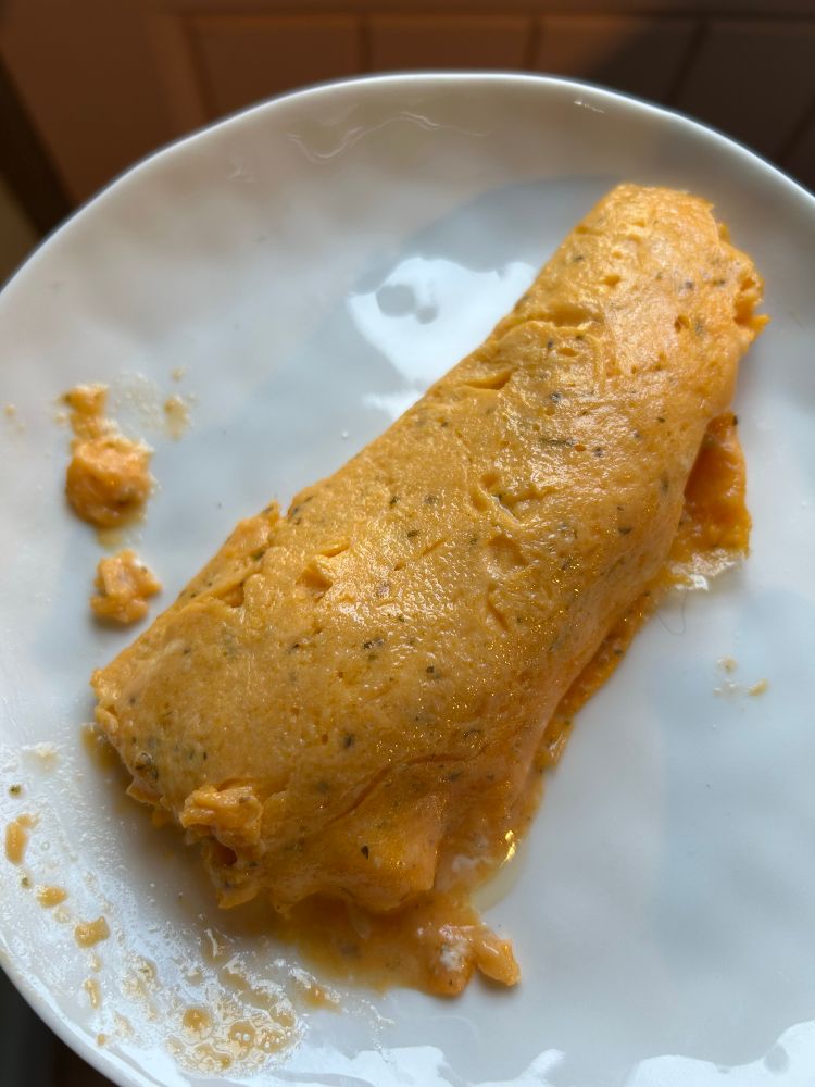 Partial eaten French style omelette on a plate. Very orange because we like our yolks that way. You can see a small bit of the soft gooey bits of the egg from the end that’s been eaten from 