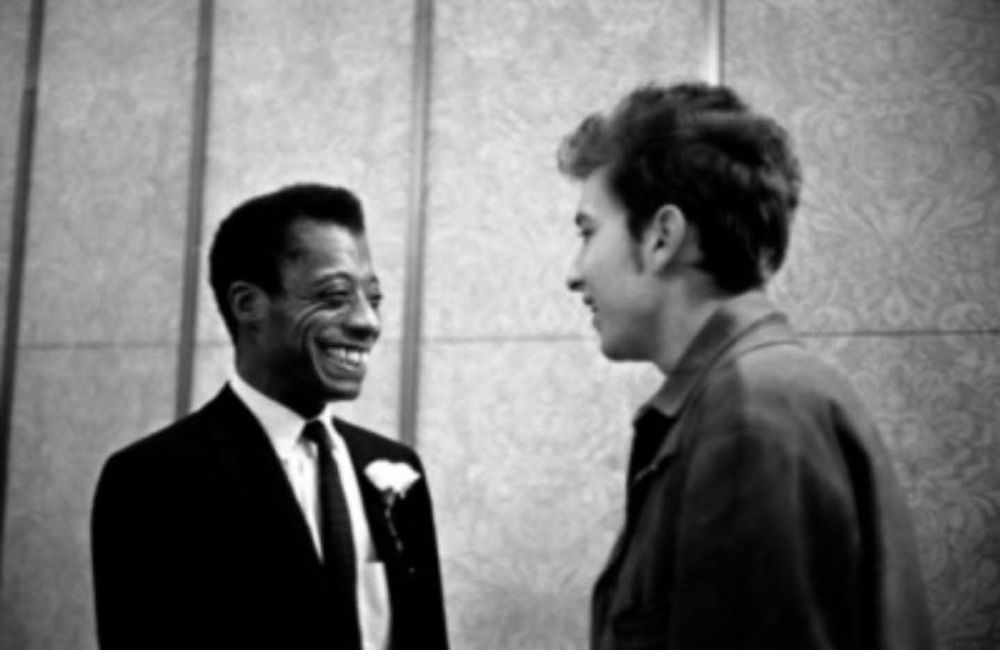 Standing and smiling James Baldwin wearing a suit and tie with a boutonniere talking to a smiling Bob Dylan.