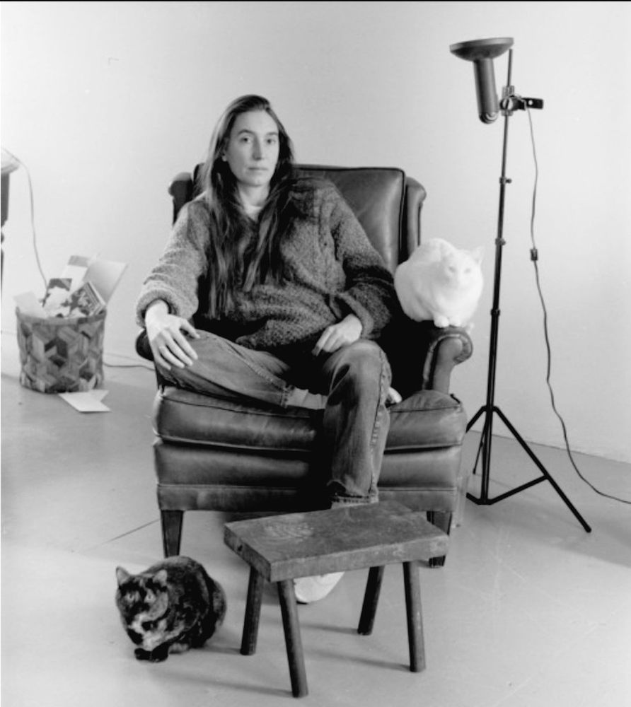 Jenny Holzer sitting in a chair foot tucked under knee, with a table in front of her, and a cat at her feet. Lamp and basket on each side.