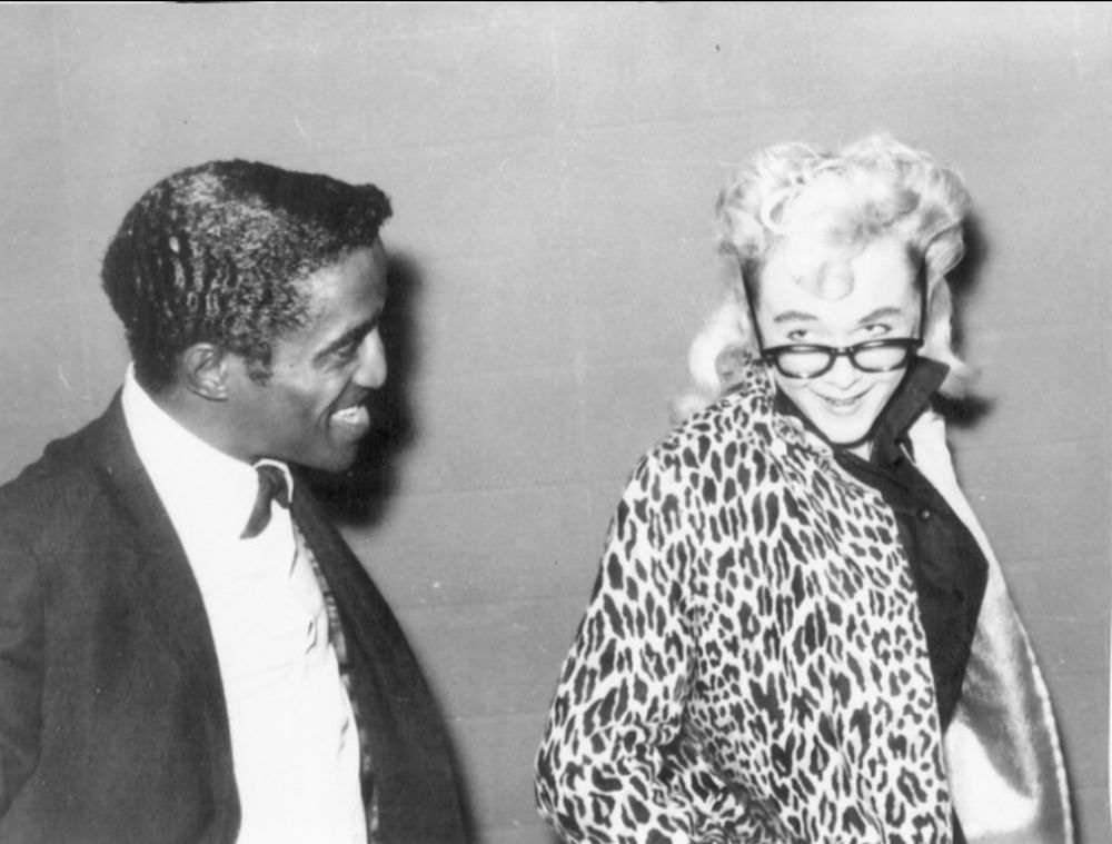 Sammy Davis Jr. smiling at a smiling Sparkle Moore wearing a leopard print jacket and peering over the top of her glasses.
