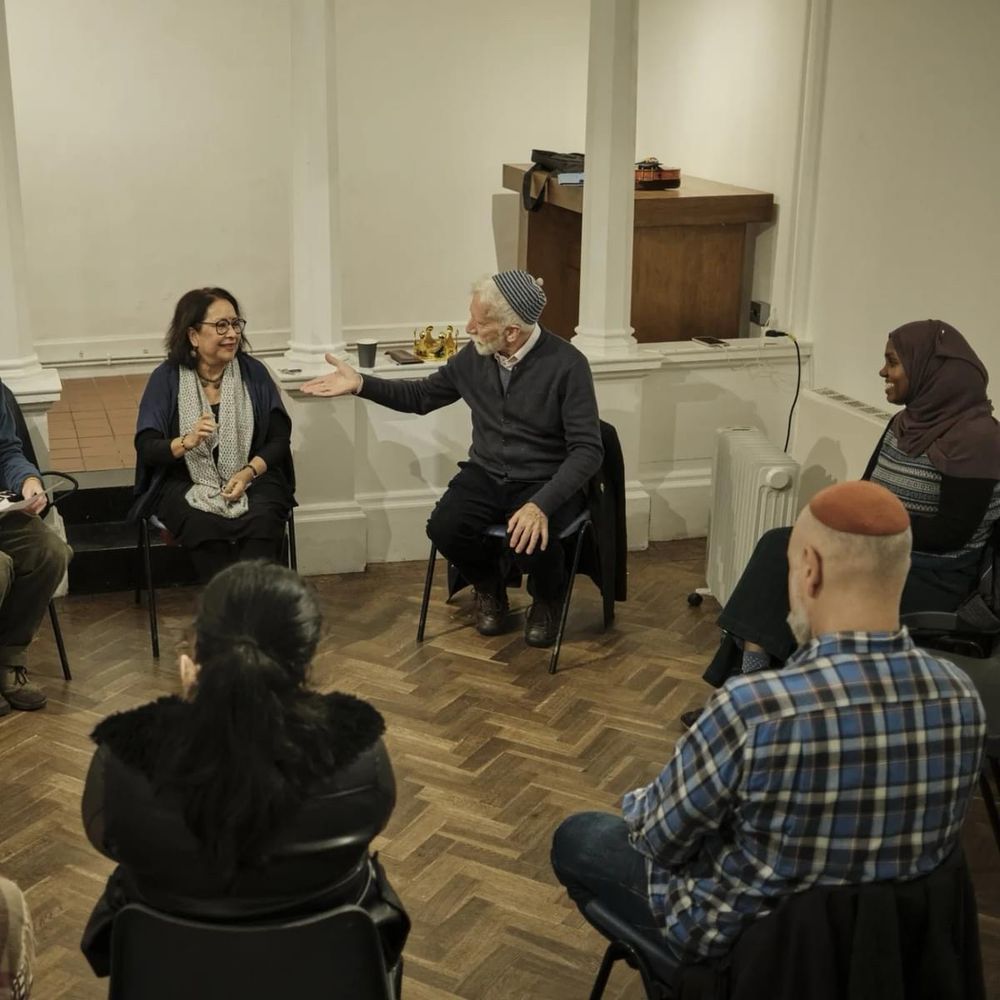 A group of people of different ethnicities sitting in a circle listening to a man with a white beard and skullcap telling a story.