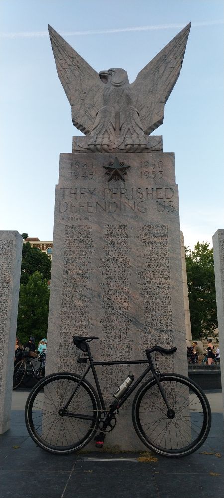 State Bicycles Core Line fixed gear bike. Black with a saddle bag, rear camera and a steel water bottle mounted on it. It is leaning on a Korean War Monument in Veterans Park in Grand Rapids, Michigan. It is a rectancular face with names and the years they died in. Also a slogan "THEY PERISHED DEFENDING US". Atop the rectangular face is a stone eagle. A dark star is in the center of the face below the eagle. It appears to be heavily patina-ed metal of some kind.

A few cyclists will about in the background.