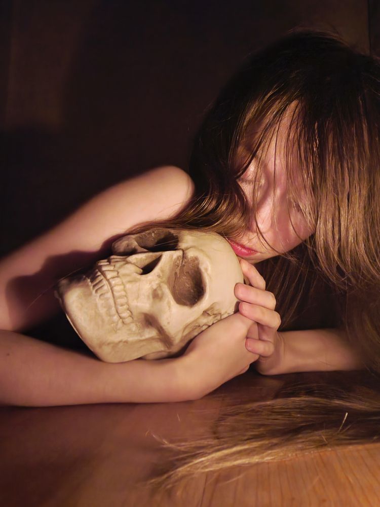 A woman is lying on the floor, facing towards a camera. Her eyes are closed and she's holding a plastic skull on her arms.